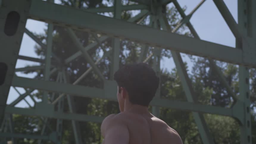 A shirtless man jogs past cars on a steel beam bridge on sunny day