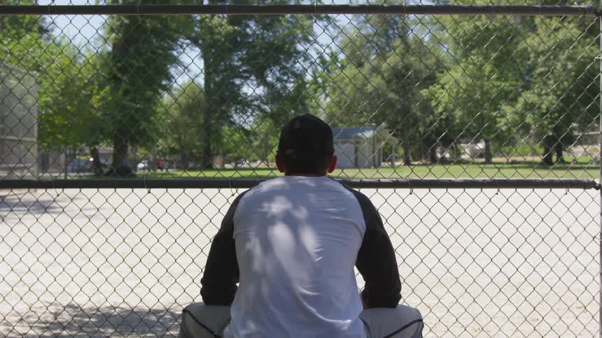 A man sits alone on a bench facing the infield and pounds his fist into his glove