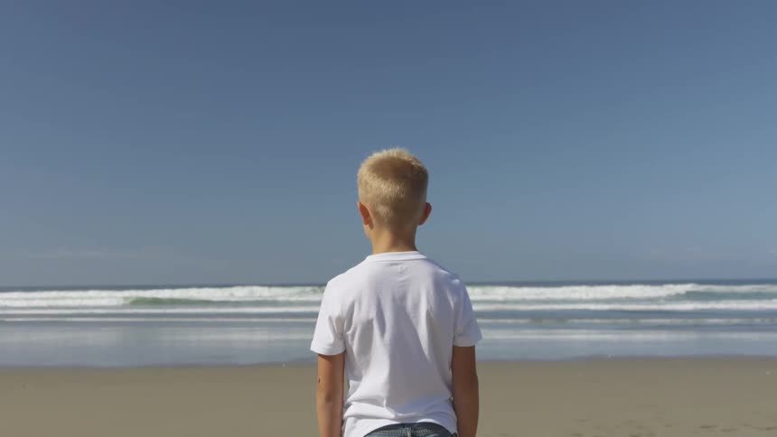 A boy standing on a beach looking at the ocean
