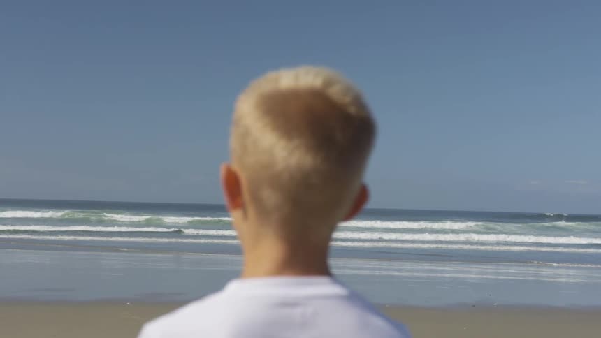 A boy standing on a beach looking out over the waves