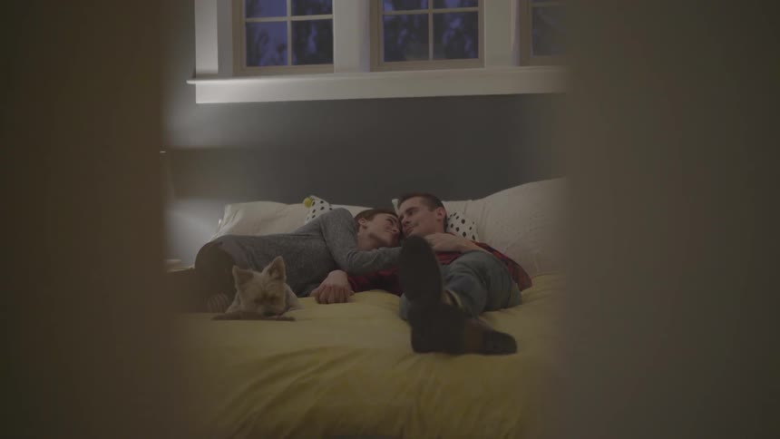 Man and woman lies down on bed with dog in bedroom at dusk