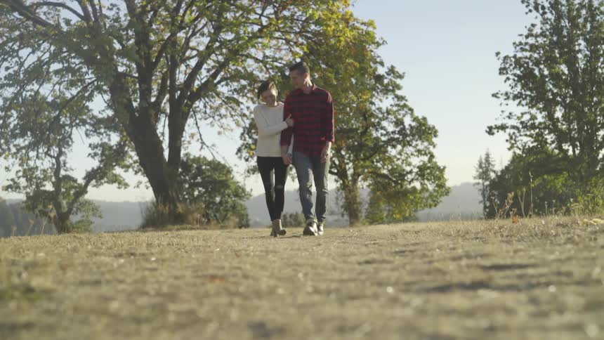 A couple walks hand in hand across a grass field heading away from the trees on a sunny day