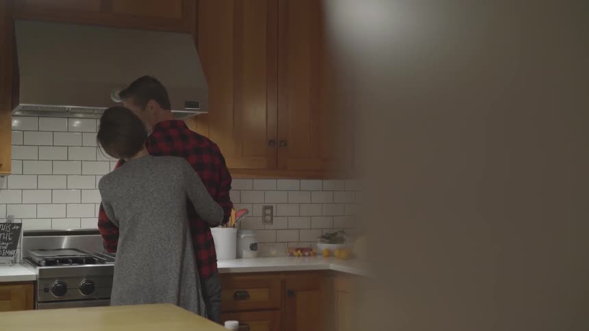 A white woman hugs a man in a plaid shirt while he cooks in a kitchen
