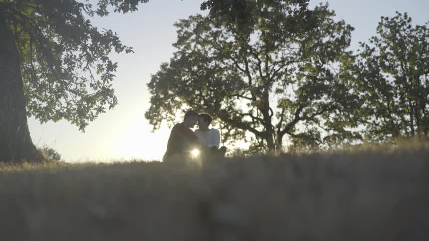 A couple sits on a grassy hillside touching foreheads