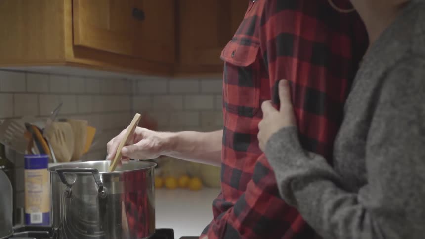 Woman hugs man's arm as he stirs pot in kitchen