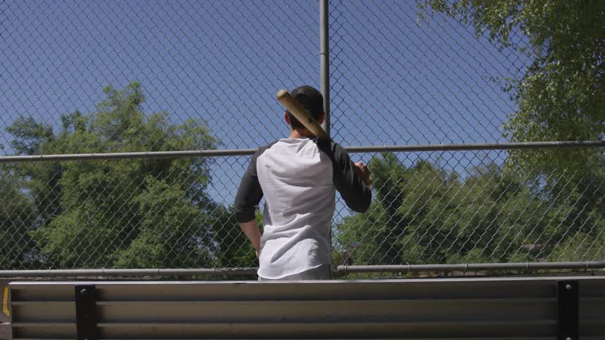 A man stands before a chain link fence with a bat on his shoulder