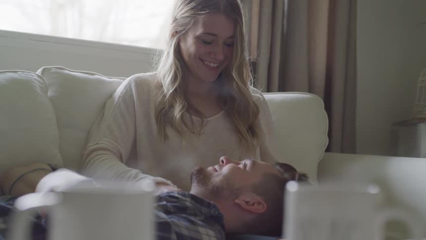 A man lays his head on a woman's lap as they sit on a couch with steaming coffee in the foreground