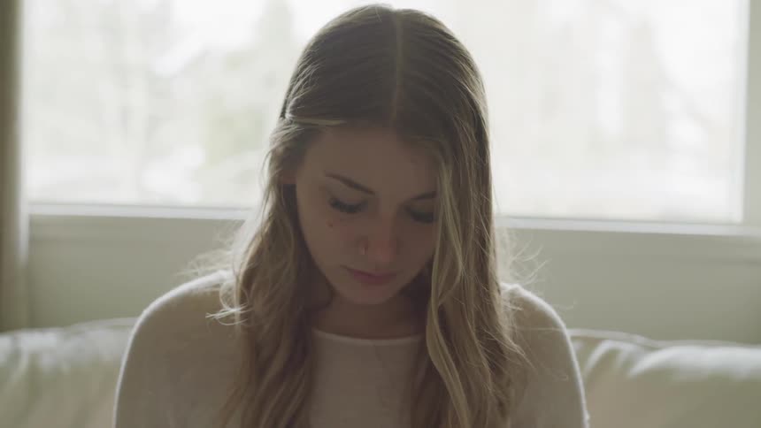 A sad looking woman sits in front of a window on a sofa
