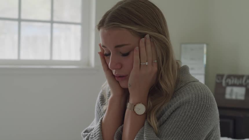 A woman sits on her bed and rubs her face with both hands