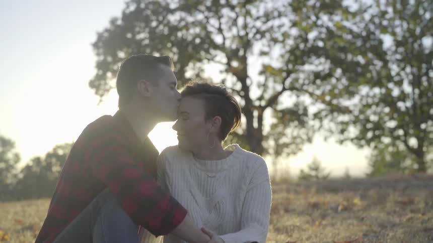 A man and a woman are sitting on a grassy hill while the man kisses the forehead of the woman