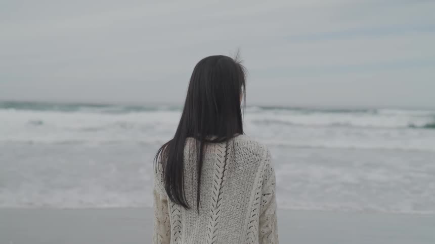 Woman stands on the beach looking at the sea whose waves crush on shore