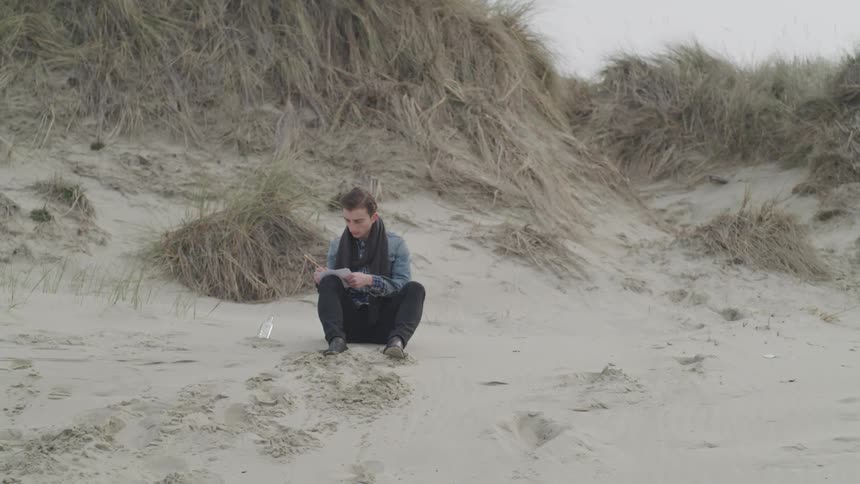 A man crouches down in sand and reads a letter from a bottle