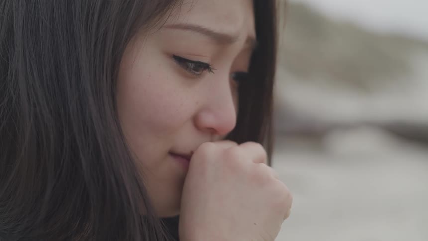 A young lady with a sad face sits with her hand covering her mouth then removes her hand and looks down