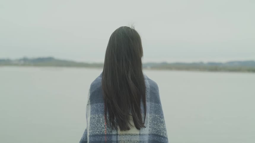 A woman stands near a lake with a blanket around her shoulders