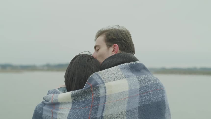 A man and a woman sit under a blanket looking out over the water