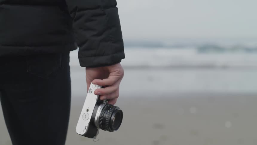 A person in black skinny jeans walking while holding a camera in their right hand