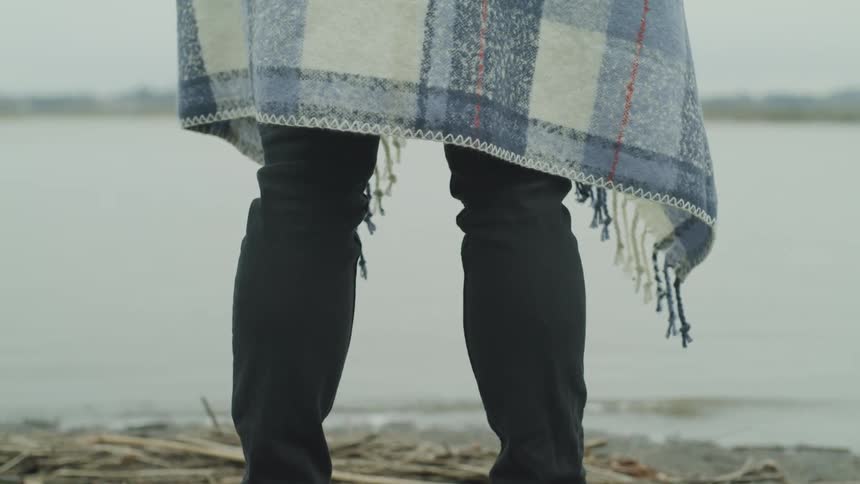 A person wrapped in a blanket stands on the shore of a lake