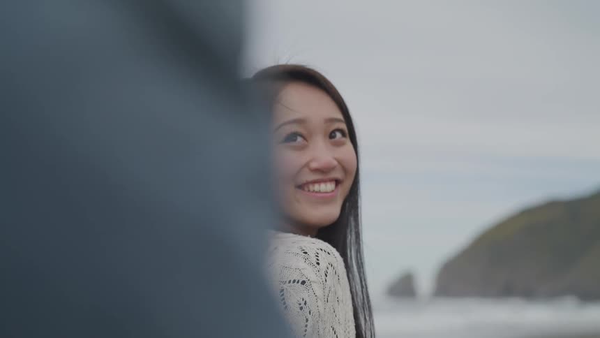 A woman stands on a beach and looks over her shoulder then smiles at someone behind her