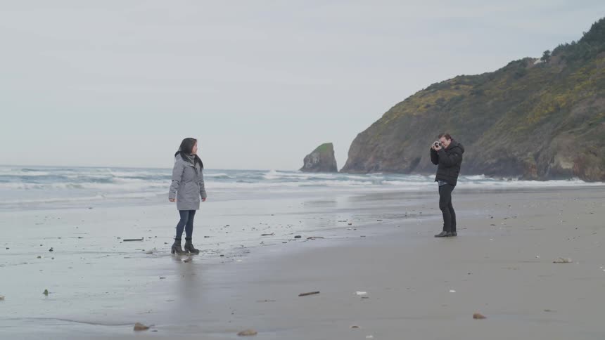 A man takes a photo of a woman as the two of them stand on a beach