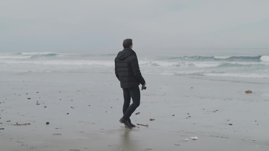 A man in a black coat walks on a beach with a camera