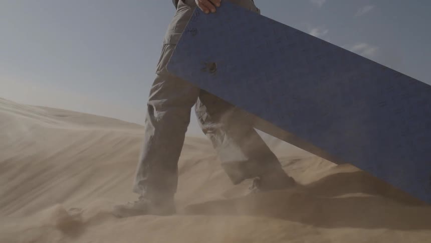 A man walks through sand and down a sand dune as he drags scrap metal behind him