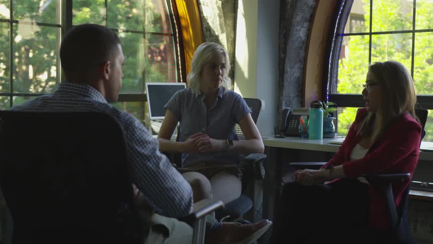 Two women and a man sit on office chairs in a office with arched windows and talk