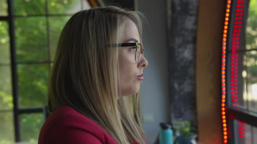 A bespectacled woman in a red blazer looks out of a window