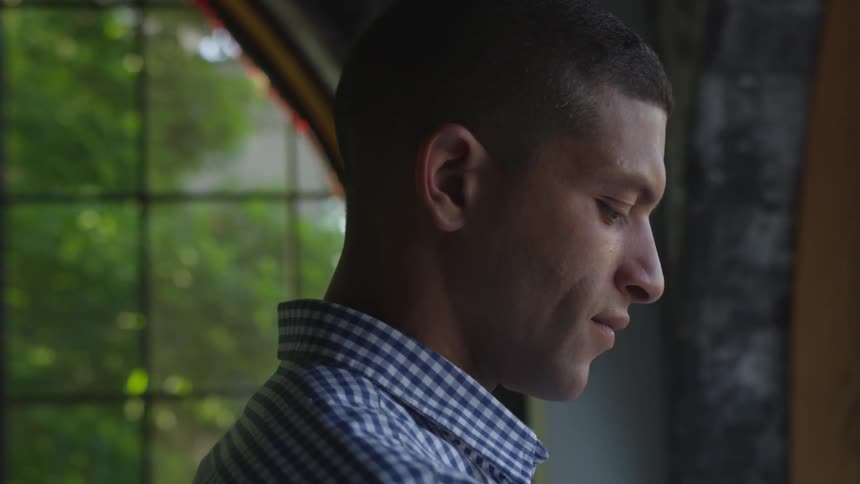 A man standing at a window looking out bends his head and looks down