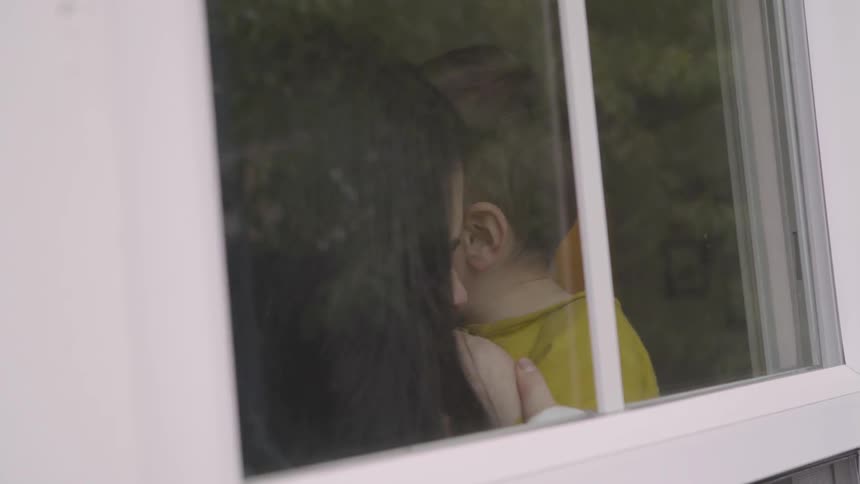 A mother plays with her child by pretending to bite her arm as they sit near a window