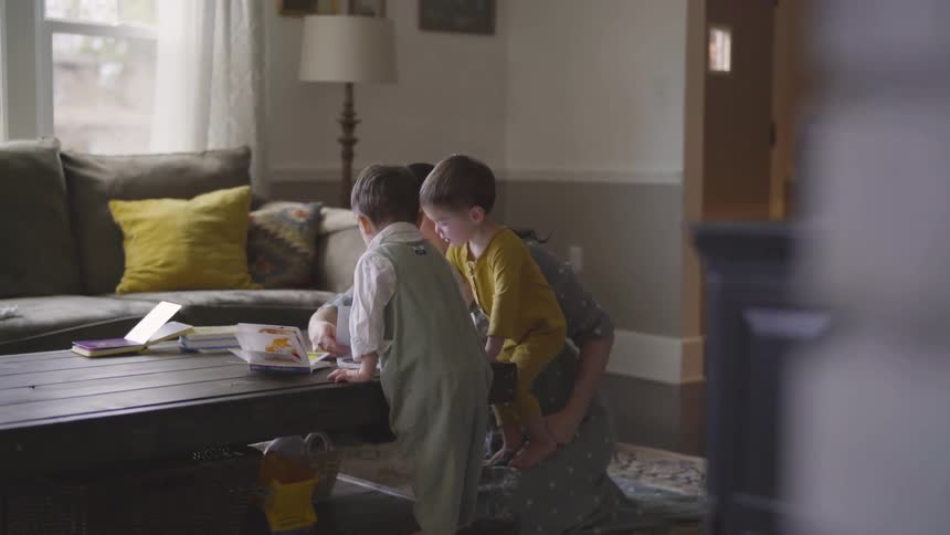 A woman shows a picture book to two boys in their living room at day