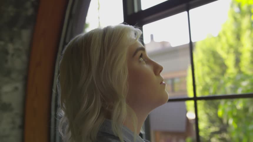A young woman stands in front of a window and glances up