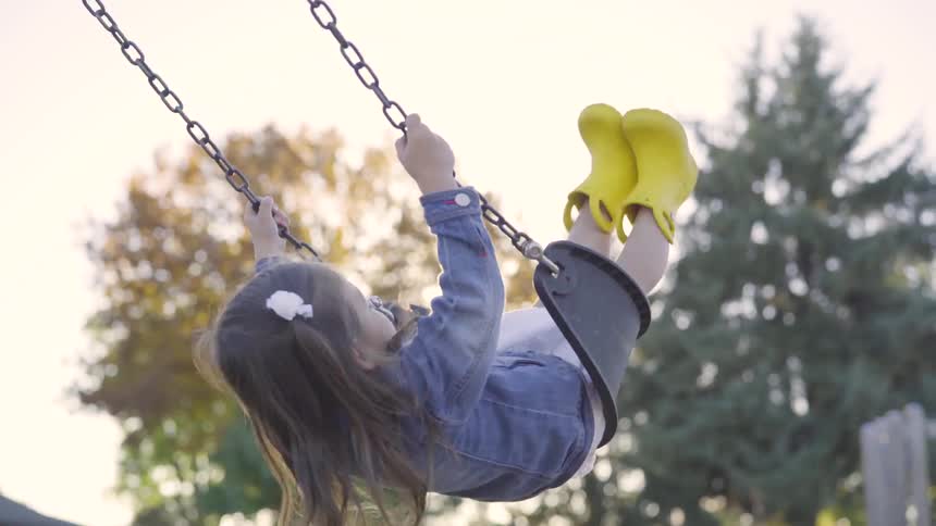 Little girl wearing yellow rain boots and denim jacket playing on swings at the park
