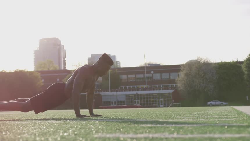 A shirtless man does push ups on the turf of an athletic field on a sunny day