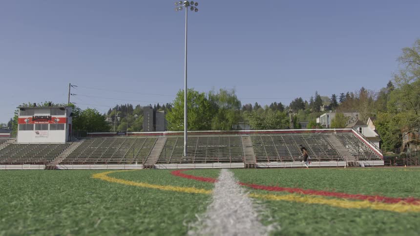 A young man in football gear runs in front of bleachers on a football field