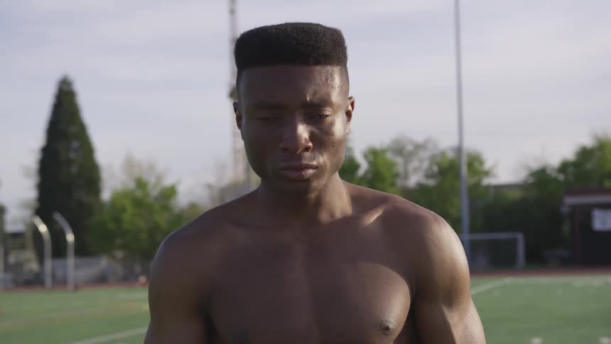 A shirtless and muscular young black man stands on an empty sports field and looks forward with poise and confidence
