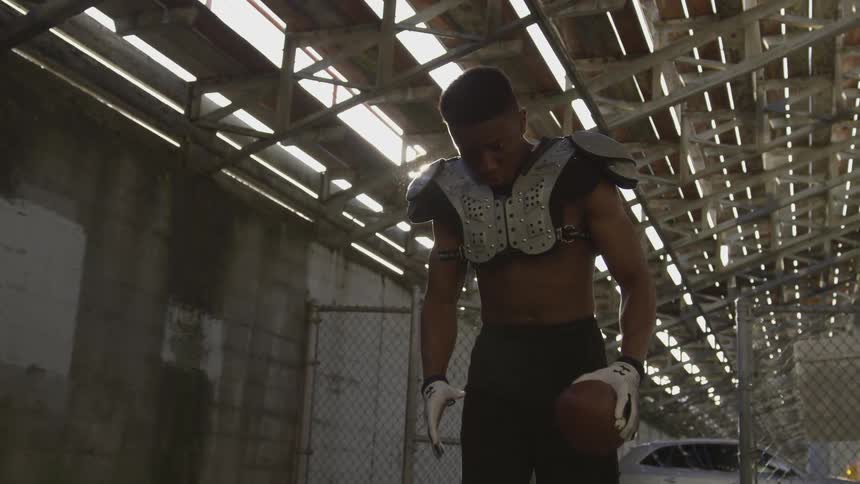 A football player wearing shorts and pads slaps the football he is holding while looking down