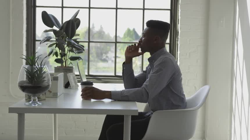 Black man sits at the table while holding plastic coffee mug and thinking indoors during daylight