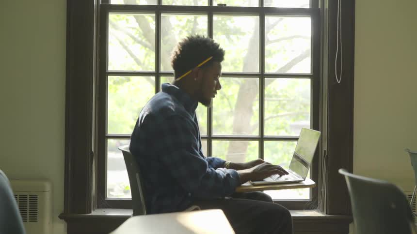 A black man sits in a classroom and types on a laptop