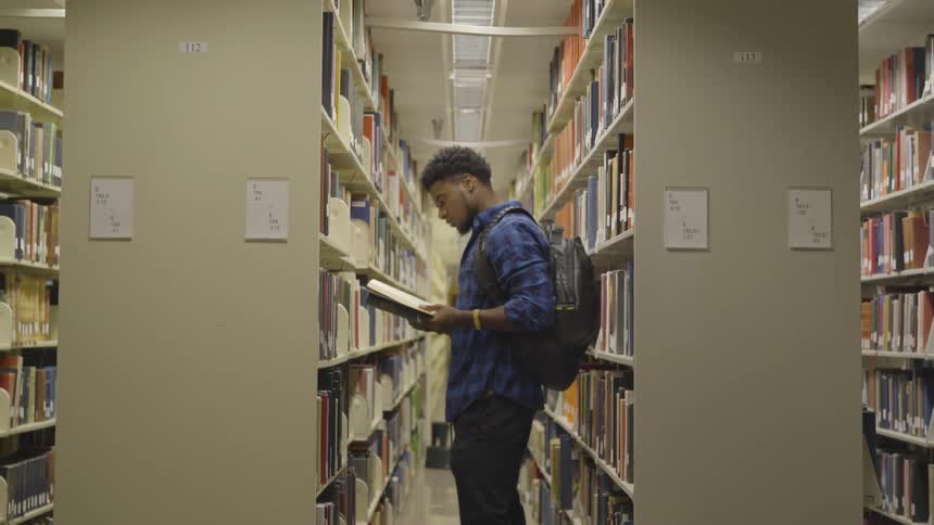 A male wearing a backpack is standing in a library searching through the books then pulls one off the shelf in front of him and starts to read