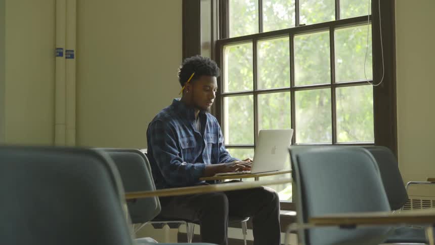 A man with a pencil behind his ear sits in a classroom typing on a laptop