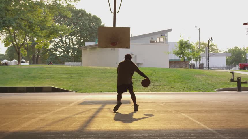 Black man wearing hoodie plays basketball on the concrete ground at sunset