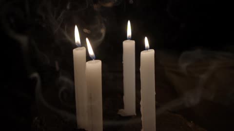Four tall candles are burning as the smoke floats around them