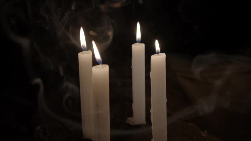 Four tall candles are burning as the smoke floats around them