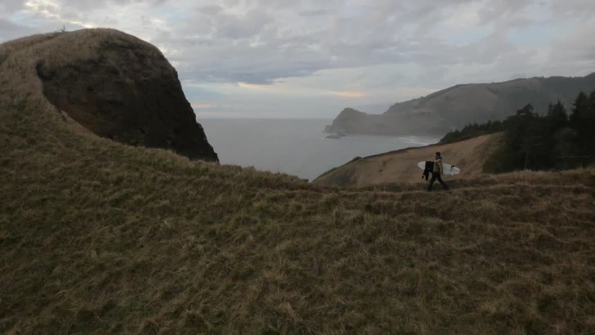 Aerial shot of a man carrying a surfboard as he walks along a rocky cliff on the coast