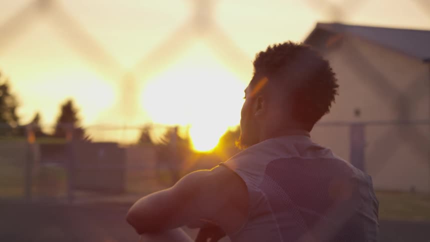 A young black man sits outside of a building in a park lined by fences and looks out contently as the sun is low in the sky