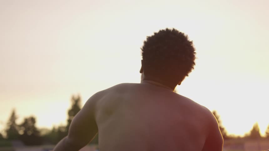 A young man with no shirt is holding onto the chain link fence his arms apart he raises and shakes his head as if as in despair