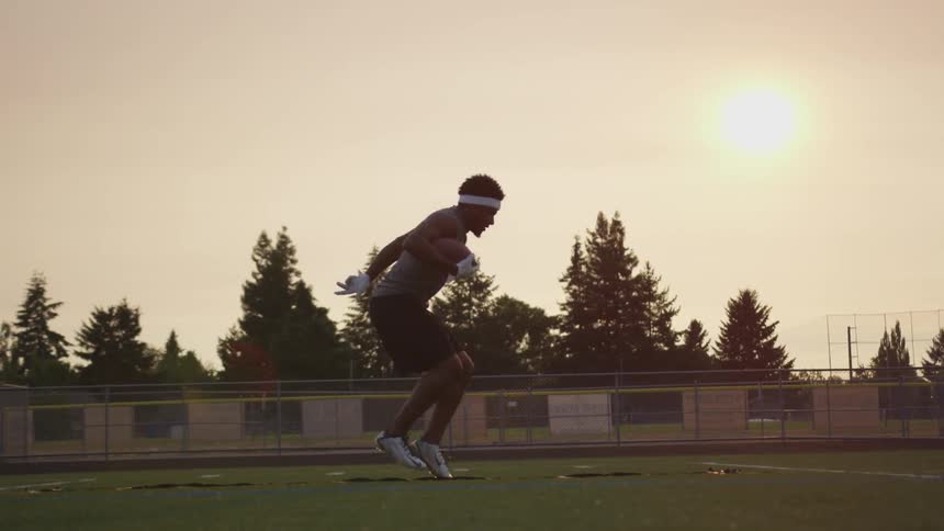 An athlete holding a football does training drills on an empty field at sunset
