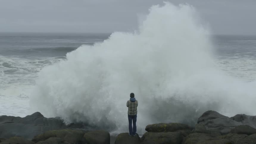 A man standing on a boulder as a large wave crashes onto the rocks before him on a cloudy day