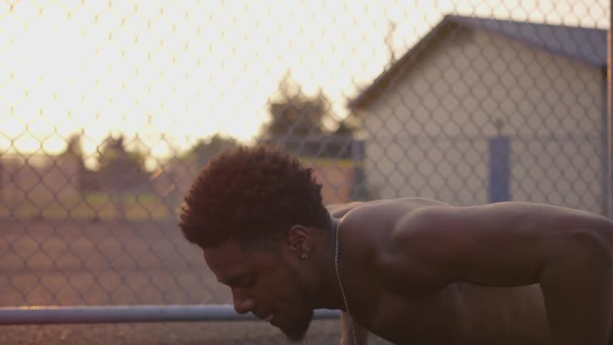 A muscular man does push ups on the ground outside near a fence