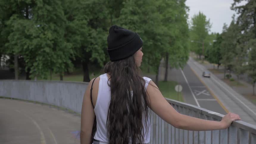 Girl wearing black wooly hat walks down on overpass by touching the banister on a cloudy day